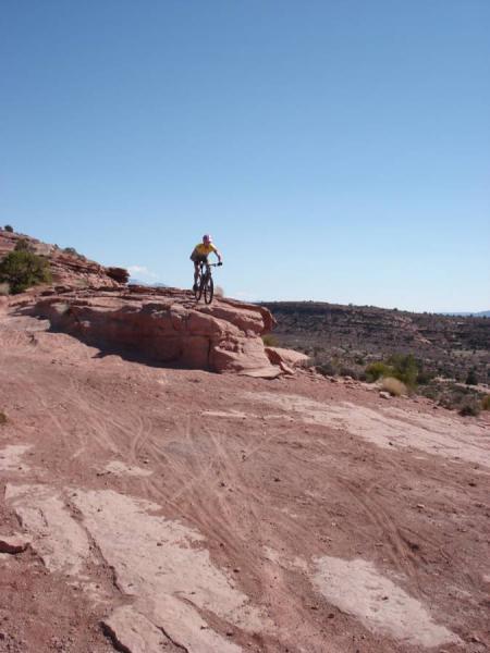 A mountain biker in yellow is riding on a rocky ledge against a clear blue sky, set in a desert landscape with red rock formations and distant hills. Porcupine Rim mountain bike trail.