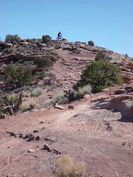 A mountain biker navigates a rocky slope under a clear blue sky, surrounded by sparse vegetation and rocky terrain. The scene captures the adventurous spirit of outdoor biking in a rugged landscape. Porcupine Rim mountain bike trail.