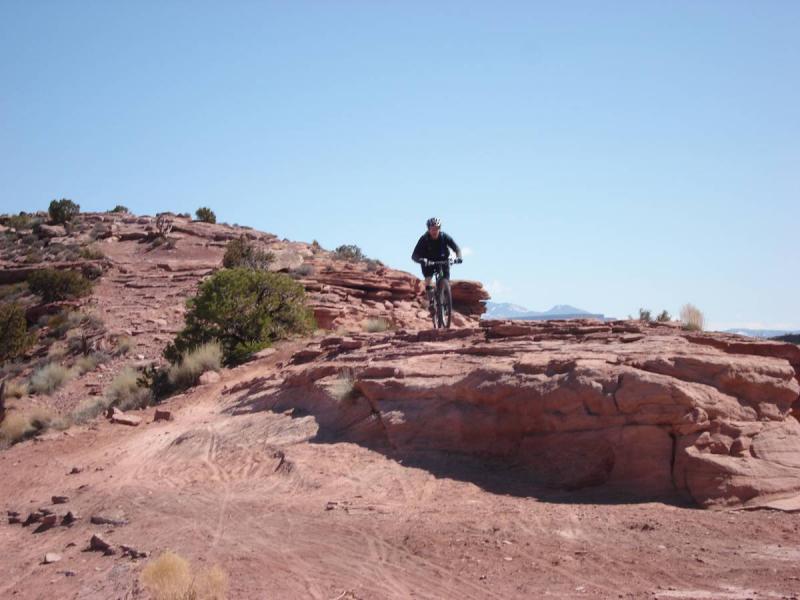 A mountain biker navigating rocky terrain under a clear blue sky, surrounded by rugged landscapes and sparse vegetation. Porcupine Rim mountain bike trail.