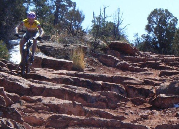 A mountain biker in a yellow shirt and helmet rides down a rocky trail surrounded by sparse vegetation and trees under a clear blue sky. Porcupine Rim mountain bike trail.