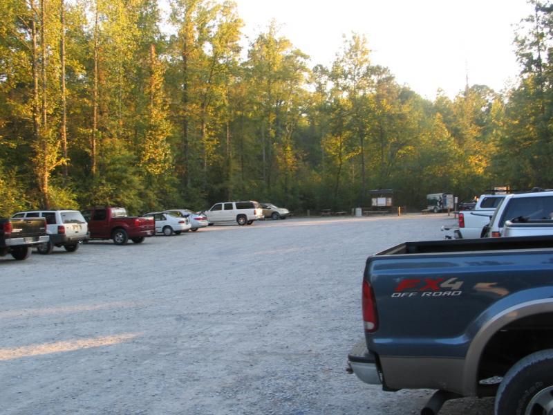 A gravel parking lot surrounded by tall trees, with several parked vehicles in various colors and sizes. The scene is illuminated by soft morning light, indicating early hours. In the background, some informative signs and a trailhead can be seen amidst the greenery. Blankets Creek mountain bike trail.