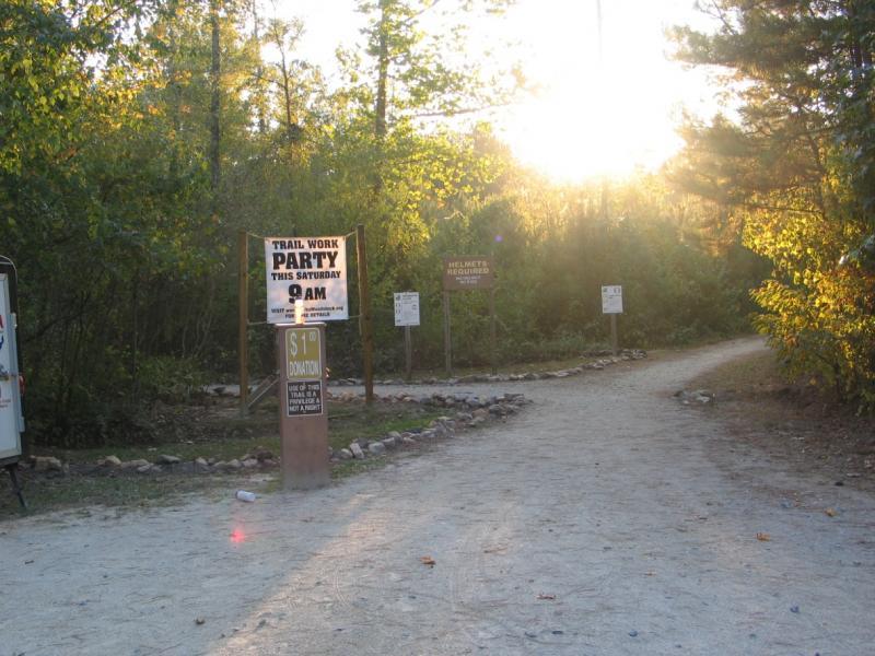 A dirt path forks in a forested area, with a large sign announcing a "Trail Work Party" scheduled for Saturday at 9 AM. The sign indicates a $1 donation is requested. There are additional signs nearby regarding safety gear, and the scene is illuminated by the warm glow of the setting sun filtering through the trees. Blankets Creek mountain bike trail.