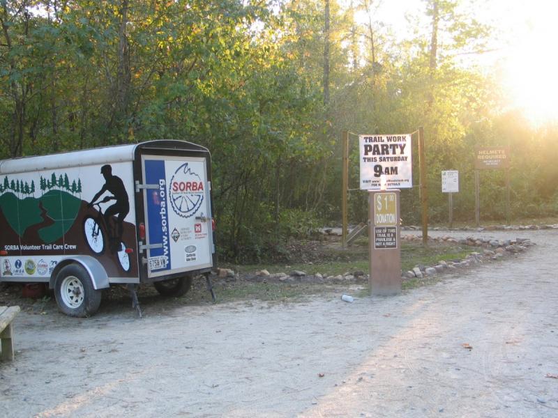 A gravel path leading into a wooded area, with a trailer displaying a biking silhouette and the text "SORBA Volunteer Trail Care Crew." There is a sign announcing a "Trail Work Party" scheduled for Saturday at 9 AM, and a donation request box nearby. The scene is illuminated by soft sunlight filtering through the trees. Blankets Creek mountain bike trail.