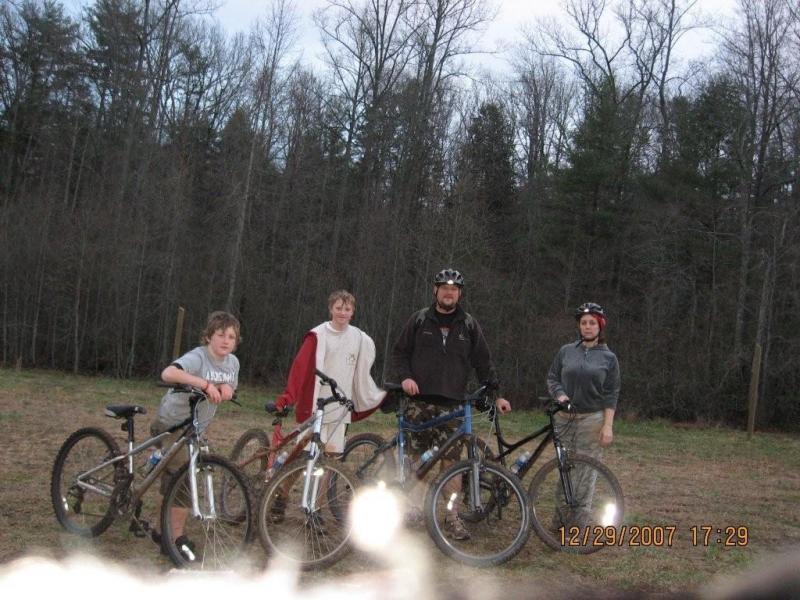 Four individuals standing on a grassy area near a wooded backdrop, each holding mountain bikes. Two boys are positioned on the left, the first wearing a gray shirt and the second in a white shirt with a red design. The third person, an adult male, is in the center wearing a dark jacket and a helmet. A fourth person, a female, stands on the right in a gray hoodie and helmet. The scene captures a casual outdoor setting, likely before a biking adventure. Bull / Jake Mountain mountain bike trail.
