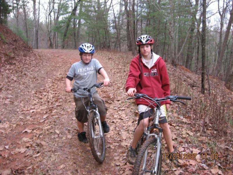 Two boys riding mountain bikes on a dirt trail surrounded by trees. One boy wears a gray t-shirt and black shorts and is pushing his bike while the other wears a red hoodie and shorts, holding the handlebars of his bike. The ground is covered with fallen leaves. Bull / Jake Mountain mountain bike trail.