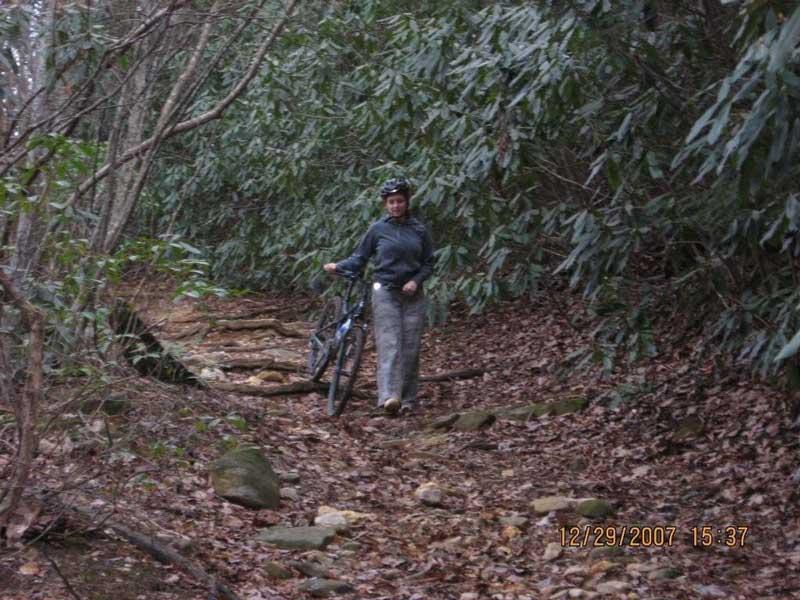 A person wearing a helmet and casual clothing walks along a narrow, rocky trail surrounded by dense greenery and fallen leaves. They are holding a bicycle while navigating the winding path through a natural setting. Bull / Jake Mountain mountain bike trail.