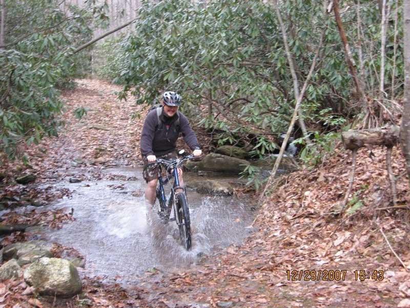 A mountain biker rides through a muddy trail with water splashing around him, surrounded by dense greenery and fallen leaves. Bull / Jake Mountain mountain bike trail.