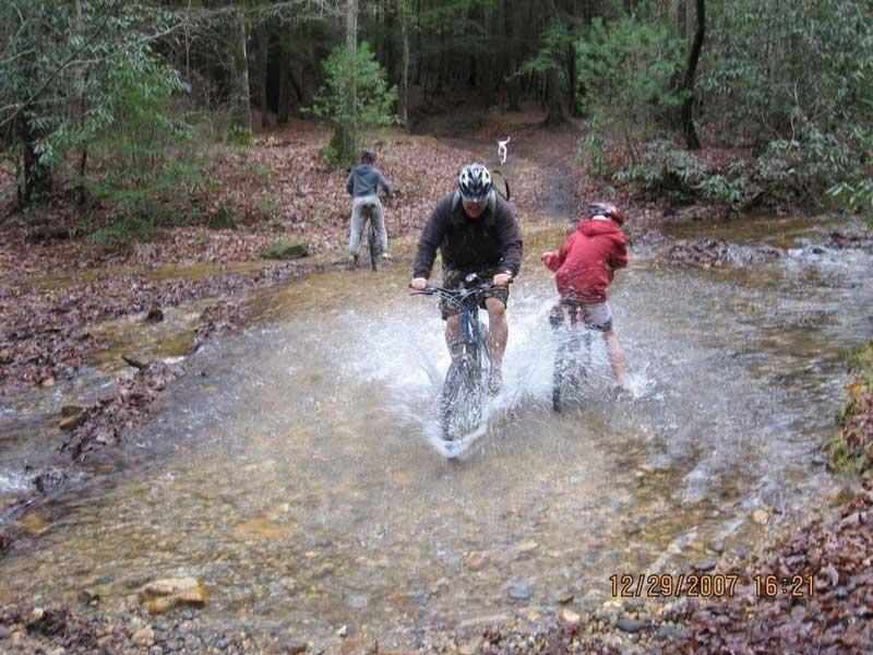 A group of mountain bikers navigating through a shallow stream in a forested area. One cyclist is pedaling through the water, creating a splash, while another has stopped beside the stream. The trail is surrounded by trees and autumn leaves cover the ground. The image captures the adventurous spirit of outdoor biking in nature. Bull / Jake Mountain mountain bike trail.