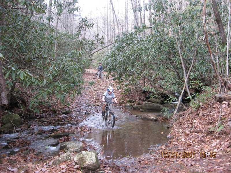 Two mountain bikers navigating a rocky, water-filled path through a forested area, surrounded by trees and fallen leaves. One biker is riding through a shallow stream, while the other is in the background, positioned slightly uphill. The scene conveys an adventurous outdoor setting. Bull / Jake Mountain mountain bike trail.