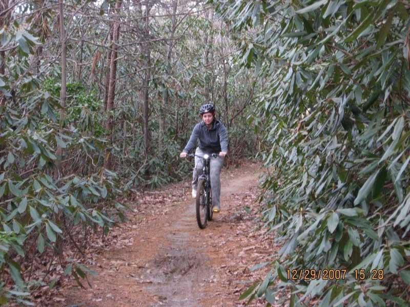 A person riding a mountain bike on a dirt trail surrounded by dense greenery, including bushes and trees, on a cloudy day. The rider is wearing a helmet and casual clothing, and the trail appears slightly muddy and winding. Bull / Jake Mountain mountain bike trail.