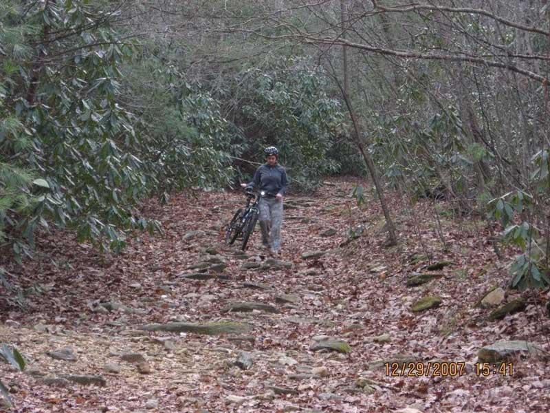 A person wearing a helmet carries a bicycle along a rocky, leaf-strewn path surrounded by trees and foliage. The scene appears to be in a forested area, likely on a hiking or biking trail. Bull / Jake Mountain mountain bike trail.