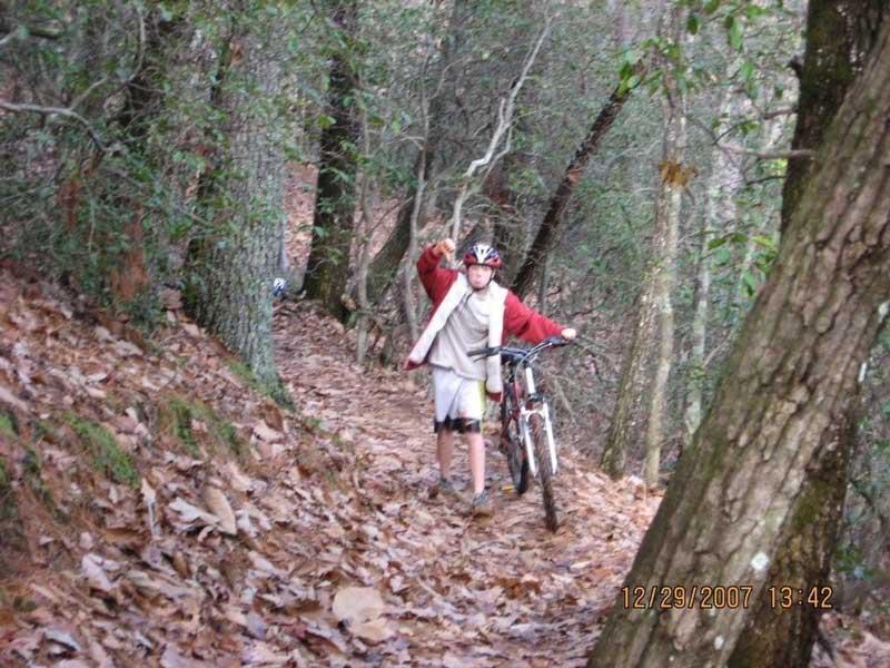 A child wearing a helmet and casual clothing is walking along a leaf-covered trail in the woods while pushing a bicycle. The surrounding trees are dense, and the path is winding, indicating a nature setting. The child is smiling and gesturing with one hand. Bull / Jake Mountain mountain bike trail.