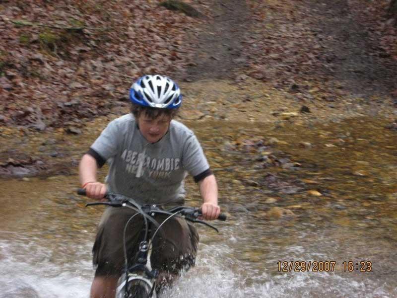 A child wearing a blue helmet and a gray T-shirt rides a bicycle through a shallow stream, splashing water as they pedal. Surrounding them are fallen leaves and a natural, wooded landscape. Bull / Jake Mountain mountain bike trail.