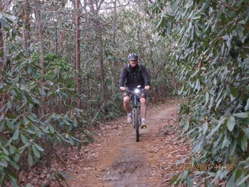 A person riding a mountain bike on a narrow trail surrounded by dense greenery and trees. The cyclist is wearing a helmet and a jacket, with a light on the front of the bike. The scene conveys a sense of outdoor adventure in a natural setting. Bull / Jake Mountain mountain bike trail.