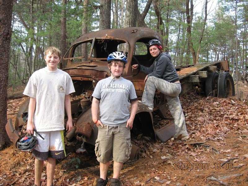 Three children are posing near an old, rusted truck in a forested area. The boys are wearing casual clothes and holding helmets, while one girl is sitting on the truck's remnants. The ground is covered with fallen leaves, and the scene is surrounded by tall trees. Bull / Jake Mountain mountain bike trail.