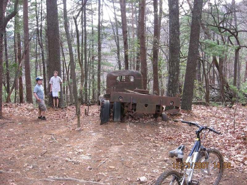 Two boys stand in a wooded area near an old, rusted truck partially obscured by trees and fallen leaves. One boy is wearing a helmet and looking at the camera, while the other stands beside a tree. A bicycle rests in the foreground, adding to the outdoor scene. Bull / Jake Mountain mountain bike trail.