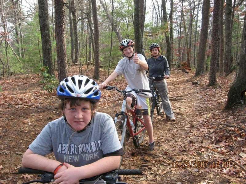 Three people, all wearing helmets, pose with mountain bikes on a forest trail covered in autumn leaves. The foreground features a young boy looking directly at the camera, while another boy gives a thumbs-up in the middle ground. A third person stands in the background, slightly behind the bikes, creating a sense of camaraderie in an outdoor setting. Bull / Jake Mountain mountain bike trail.