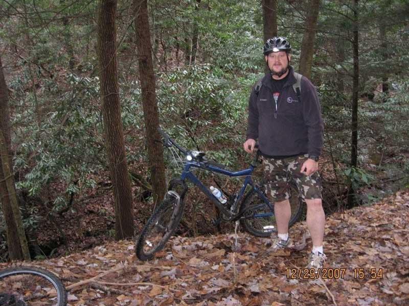 A man in a black helmet stands beside a blue mountain bike on a forest trail covered with leaves. He wears a dark sweatshirt and camouflage shorts, with a backpack on his back. The background features tall trees and greenery, indicating a natural outdoor setting. Bull / Jake Mountain mountain bike trail.