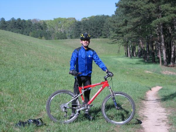 A person in a blue jacket and black pants stands beside a red mountain bike on a grassy trail, surrounded by trees and rolling hills on a sunny day. Fort Yargo State Park mountain bike trail.