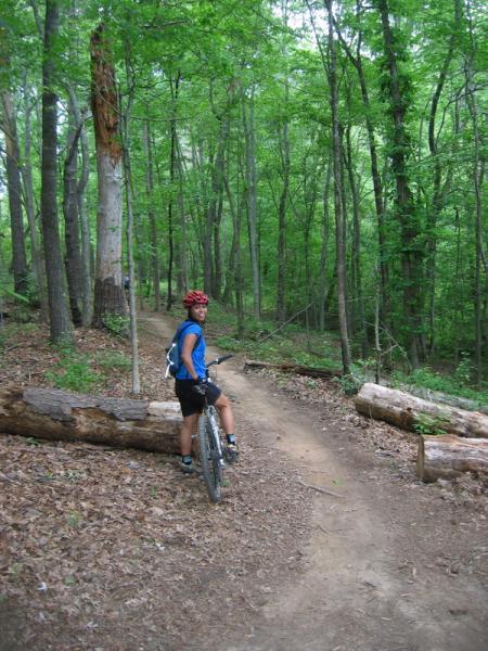 A person wearing a blue shirt and red helmet stands beside a mountain bike on a dirt trail in a lush green forest. The trail winds through trees and logs, indicating a natural, outdoor environment. Fort Yargo State Park mountain bike trail.