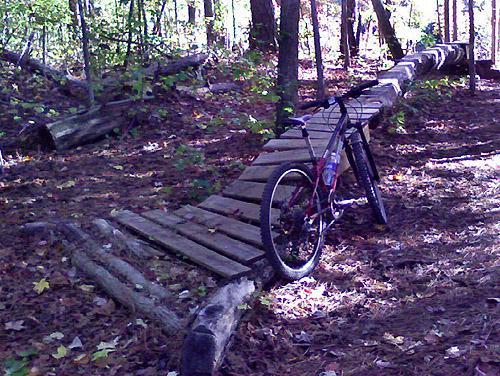A mountain bike leaning against a wooden bridge surrounded by trees and fallen leaves in a forested area. Big Creek mountain bike trail.