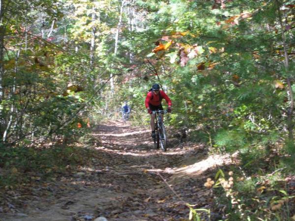 A mountain biker dressed in a red jacket rides along a dirt trail surrounded by trees and autumn foliage. A second cyclist can be seen in the background, navigating the winding path through a serene forest setting. Bent Creek mountain bike trail.