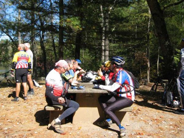 A group of cyclists taking a break at a picnic table in a wooded area, enjoying snacks and drinks. Some cyclists are seated at the table while others are standing nearby, with bicycles parked in the background. The setting features tall trees and a peaceful outdoor environment. Bent Creek mountain bike trail.