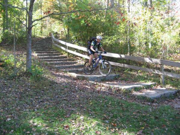 A cyclist navigating a set of stone steps while riding a mountain bike on a wooded trail, surrounded by trees and autumn foliage. Bent Creek mountain bike trail.