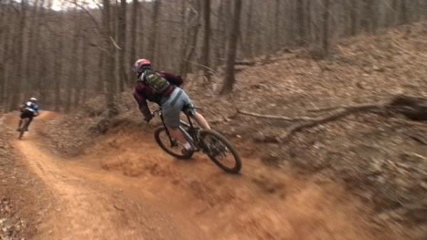 Alt text: Two mountain bikers navigate a dirt trail through a forested area. One rider leans into a turn, while the other is slightly behind on the path. The landscape features bare trees and a few patches of leaves on the ground. Bent Creek mountain bike trail.
