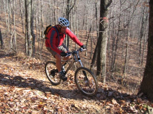 A cyclist wearing a helmet and a red jacket rides a mountain bike down a wooded trail covered with fallen leaves. Trees with bare branches surround the path, indicating it is autumn or early spring. Bent Creek mountain bike trail.