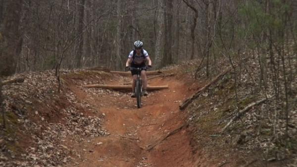 A mountain biker riding down a dirt trail in a wooded area, surrounded by trees and autumn leaves. The path has a slight incline and includes wooden obstacles, showcasing a rugged outdoor cycling environment. Bent Creek mountain bike trail.