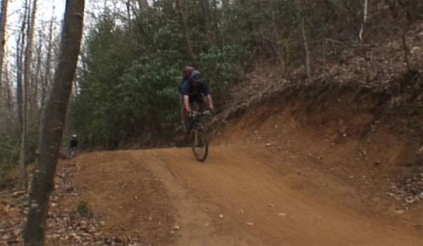 A mountain biker riding on a dirt trail in a forested area, with trees and greenery in the background. The trail is slightly uphill, and another cyclist can be seen in the distance. Bent Creek mountain bike trail.
