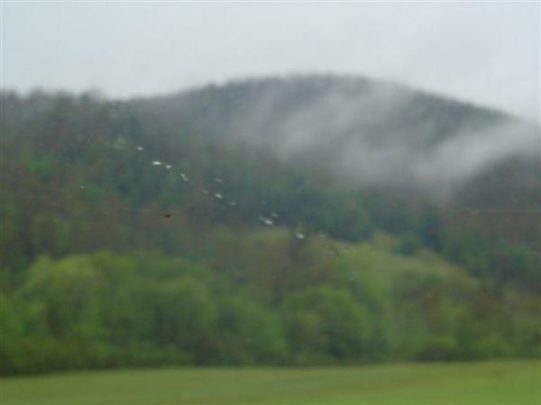 A misty landscape featuring rolling hills covered with trees, partially obscured by low clouds. The foreground shows a green grassy area with blurred raindrops on a window, suggesting rainy weather. Bent Creek mountain bike trail.