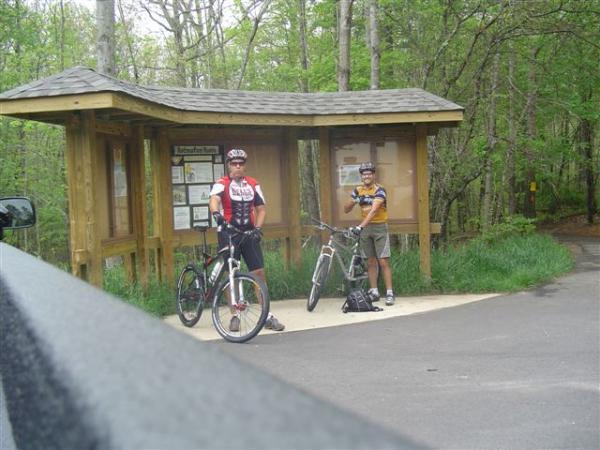 Two cyclists are standing next to their bikes near a wooden information kiosk in a forested area. The kiosk displays maps and information about biking trails. One cyclist is wearing a red and black jersey, while the other is in a multicolored jersey. The background features lush green trees typical of a wooded environment. Bent Creek mountain bike trail.