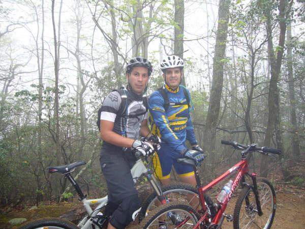 Two mountain bikers posing together on a forested trail. They are wearing helmets and biking gear, with one rider on a light-colored bike and the other on a red bike. The background features trees and a slightly foggy atmosphere, suggesting an outdoor adventure. Bent Creek mountain bike trail.