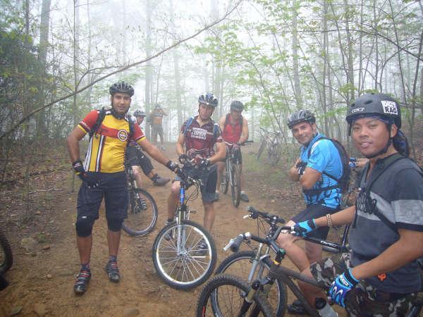 Group of mountain bikers posing for a photo on a foggy trail, surrounded by trees. They are wearing helmets and cycling gear, with several bikes visible. The atmosphere appears adventurous and the visibility is low due to mist. Bent Creek mountain bike trail.
