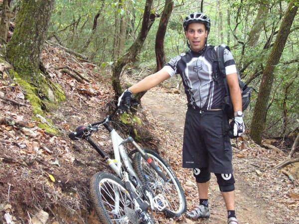 A mountain biker in athletic gear stands next to a bicycle on a dirt trail surrounded by trees. The biker is wearing a helmet, gloves, and a short-sleeved jersey with a design, and appears to be taking a break on a wooded path. Bent Creek mountain bike trail.