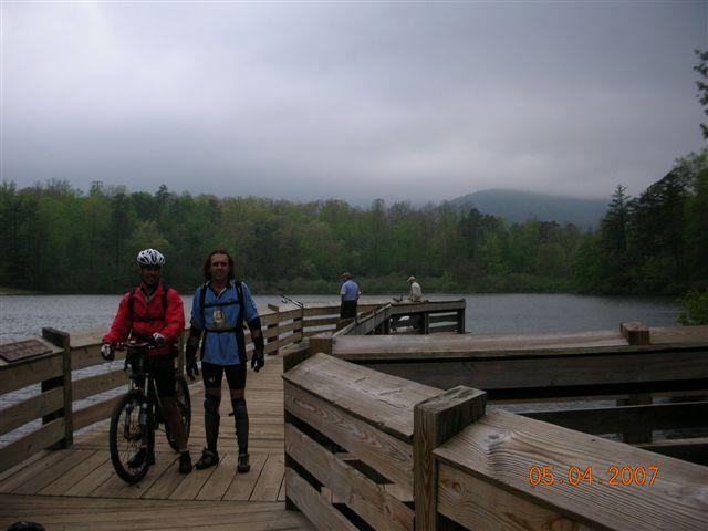 Two cyclists stand on a wooden walkway overlooking a serene lake, surrounded by lush green trees and misty mountains in the background. The sky appears cloudy, adding a tranquil atmosphere to the scene. One cyclist is dressed in a red jacket, while the other wears a blue shirt and cycling shorts. In the distance, a few people are enjoying the view from the lake's edge. Bent Creek mountain bike trail.