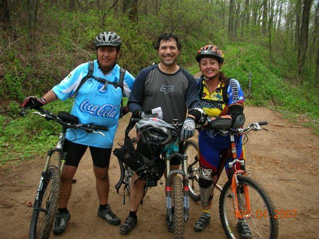 Three mountain bikers stand together on a dirt trail surrounded by green foliage. Two men wear helmets and cycling gear, while a woman in colorful attire holds her bike. They are smiling and appear to be enjoying a break during their ride. The image is dated October 4, 2007. Bent Creek mountain bike trail.