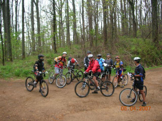 A group of mountain bikers gathered on a dirt path in a wooded area. They are wearing helmets and cycling gear, with various types of mountain bikes. The background features tall trees and green foliage, indicating a spring or summer season. The image captures a moment of camaraderie among the cyclists. Bent Creek mountain bike trail.