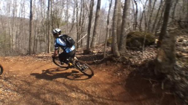 A mountain biker skillfully navigating a winding dirt trail in a forested area, with trees in the background and rocky terrain alongside the path. DuPont State Recreational Forest mountain bike trail.