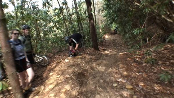 A group of three cyclists is standing near a dirt trail surrounded by dense greenery. One cyclist in the foreground is crouched down, possibly adjusting their bike, while the other two stand nearby in casual cycling attire. The trail is narrow and bordered by trees, indicating a natural, outdoor environment. DuPont State Recreational Forest mountain bike trail.