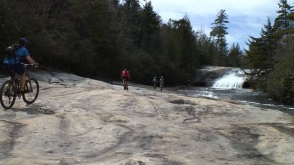 A group of three people cycling and walking along a rocky area near a waterfall, surrounded by lush trees and a clear blue sky. DuPont State Forest mountain bike trail.