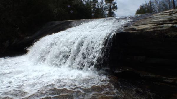 A waterfall cascading over a rocky ledge, surrounded by trees. The water flows vigorously, creating a mist in the air and forming a pool at the base. A person is visible in the background, adding a scale to the scene. DuPont State Recreational Forest mountain bike trail.