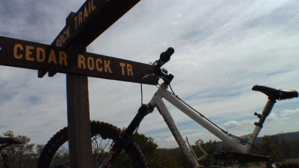 A close-up view of a mountain bike resting against a wooden trail sign that reads "CEDAR ROCK TR." The background features a cloudy sky and a hint of greenery, suggesting an outdoor setting. DuPont State Recreational Forest mountain bike trail.