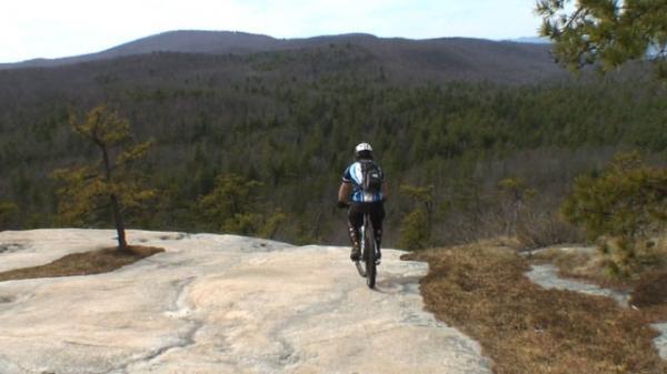 A mountain biker riding on a rocky outcrop overlooking a vast forest landscape with rolling hills in the background. The scene captures a sense of adventure and the beauty of nature. DuPont State Recreational Forest mountain bike trail.
