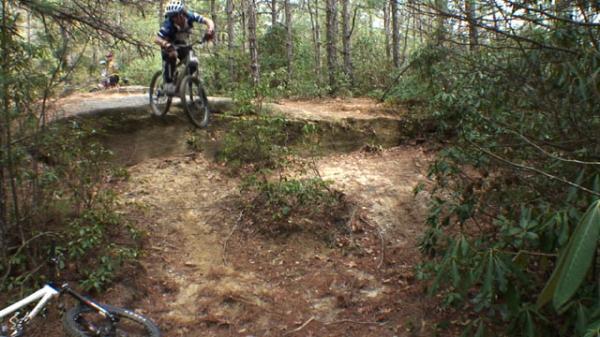 A mountain biker jumping off a rocky ledge in a forested area, surrounded by trees and underbrush. A stationary bicycle is in the foreground, while other riders can be seen in the background. DuPont State Forest mountain bike trail.