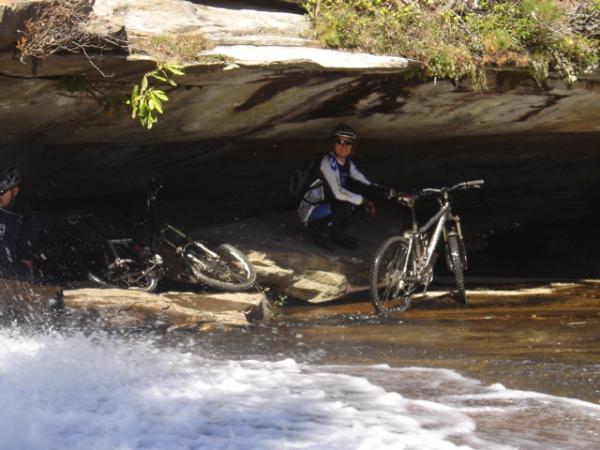 A mountain biker resting on a rock under a rock overhang, with two bicycles nearby, while water flows over the rocks in the foreground. DuPont State Forest mountain bike trail.