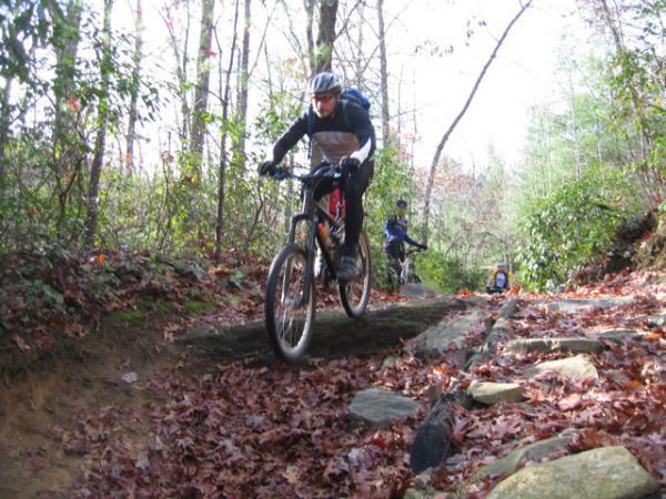 Mountain biker navigating a rocky trail covered in autumn leaves, surrounded by trees and greenery. Another cyclist is visible in the background. DuPont State Forest mountain bike trail.