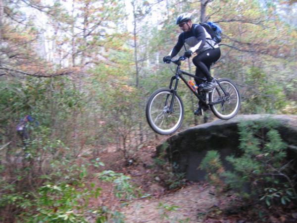 A mountain biker wearing a helmet and tight-fitting clothing is captured mid-air, jumping from a large rock in a wooded area. Surrounding the biker are trees and greenery, indicating an outdoor trail suitable for biking. DuPont State Forest mountain bike trail.
